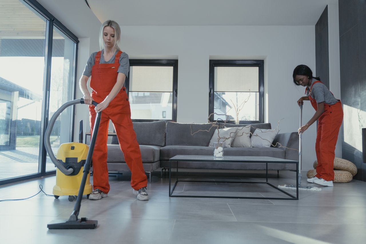 brand-02 Two women in red overalls vacuuming and sweeping a modern living room.